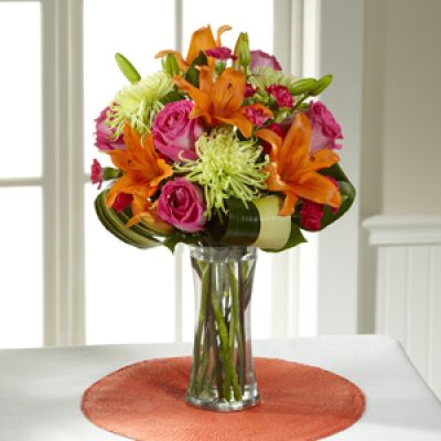 Hot pink bi-colored roses, orange Asiatic Lilies, green Fuji Chrysanthemums, hot pink mini carnations, tropical leaves, and lush greens are brought together to impress, presented in a clear glass vase. Bright, beautiful, and ready to get the party started, this bouquet creates a unique surprise to absolutely delight your recipient with each sunlit bloom.